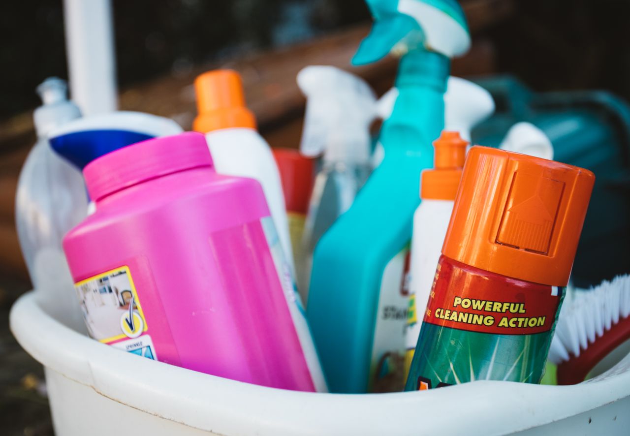 Close-up view of assorted cleaning products gathered in a bucket outdoors, highlighting a range of detergents and sprays.